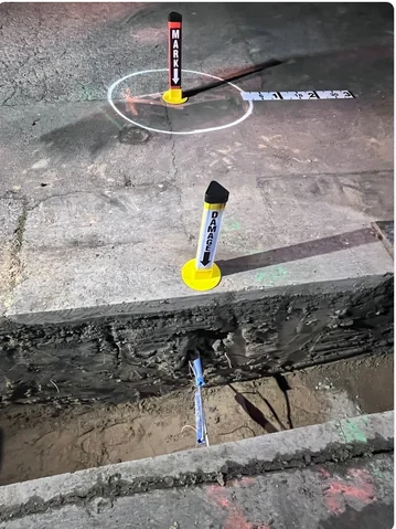 Open excavation with a damaged line exposed, marked by MARKER and DAMAGE posts with a measuring scale laid across the trench