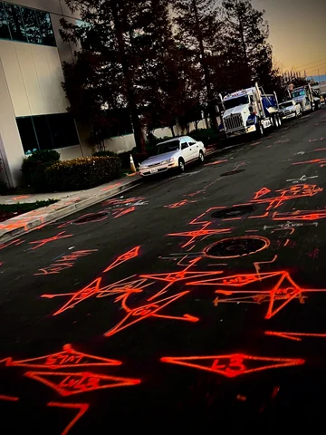 Utility locate markings painted across a street at dusk with work trucks staged alongside